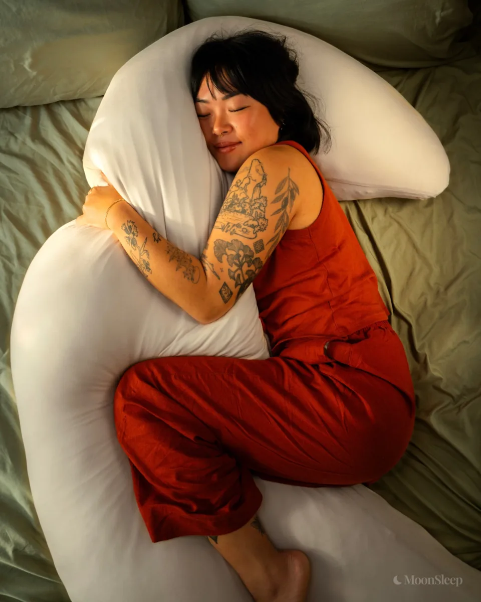 Asleep and hugging the MoonSpoon cuddle pillow on olive sheets, arms and legs wrapped around the crescent
